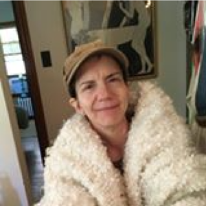 Smiling woman indoors wearing a tan cap and a fluffy cream coat.